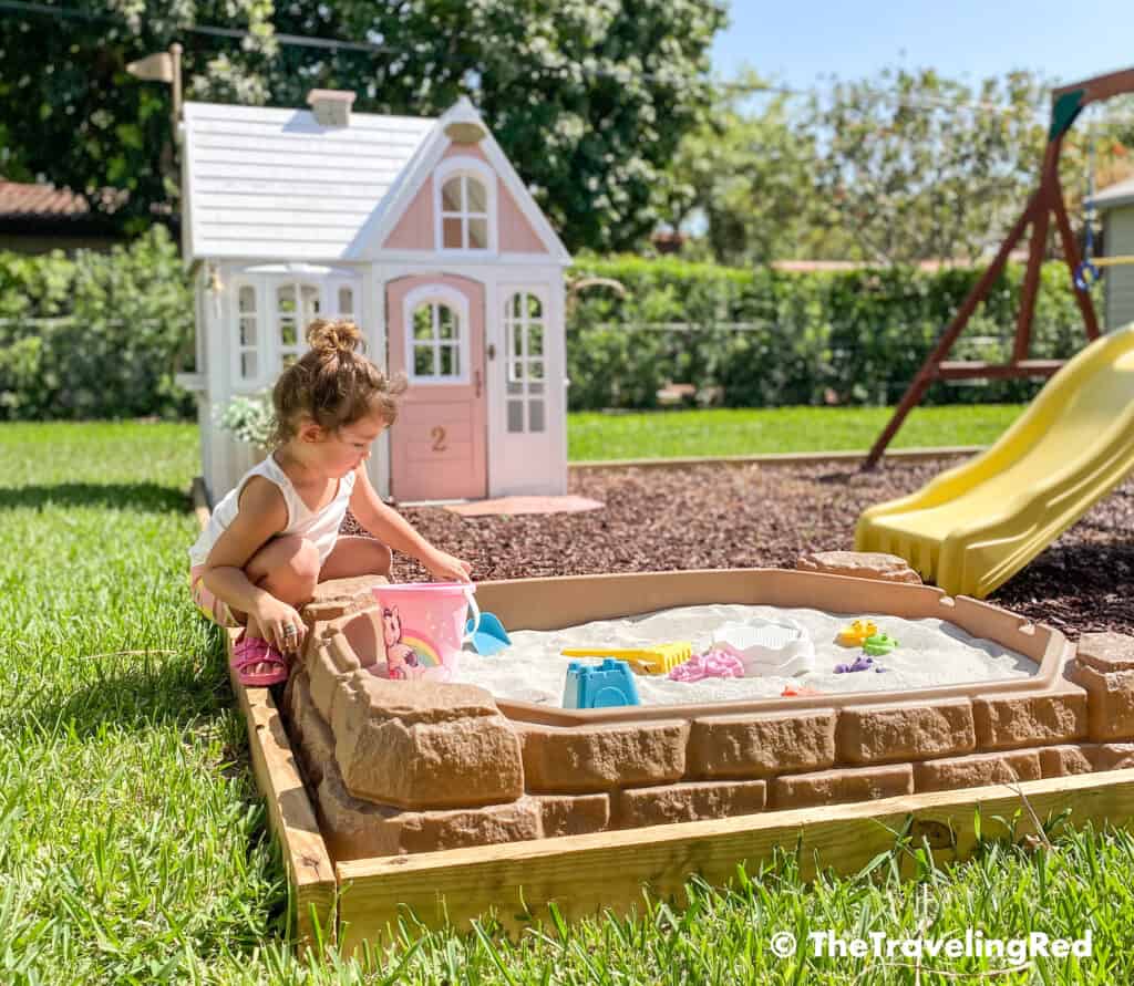How we built a custom backyard playground with rubber mulch for our swingset, playhouse and sand box. Our sandbox is filled with beach toys and she loves playing in it for hours on end. This outdoor playground is perfect little outdoor play space for your kids and it's soft on the feet.