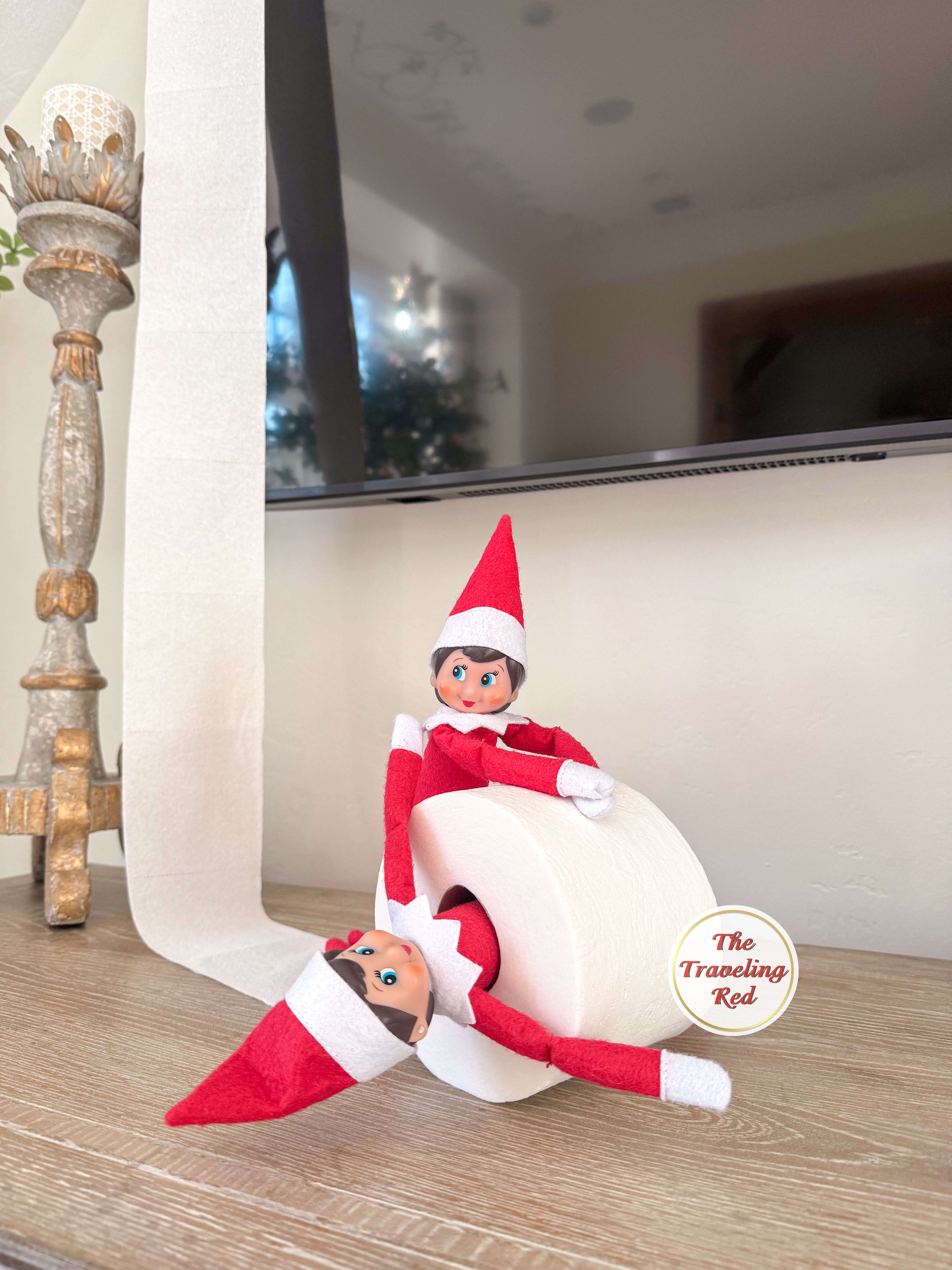 Elf on the Shelf Ideas: Elf is playing with another elf friend and a roll of toilet paper. One elf is inside the roll and the other is pushing it. Over 100 easy and quick elf on the shelf ideas that are fun. By The Traveling Red