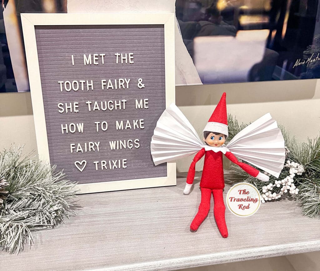 Elf on the Shelf Ideas: Elf is wearing wings made of a sheet of paper. The white sheet of paper is folded up and put on her back using clear hair ties. The message board reads, I met the tooth fairy and she taught me how to make fairy wings. This is for a day your child lost a tooth. Over 100 easy and quick elf on the shelf ideas that are fun. By The Traveling Red