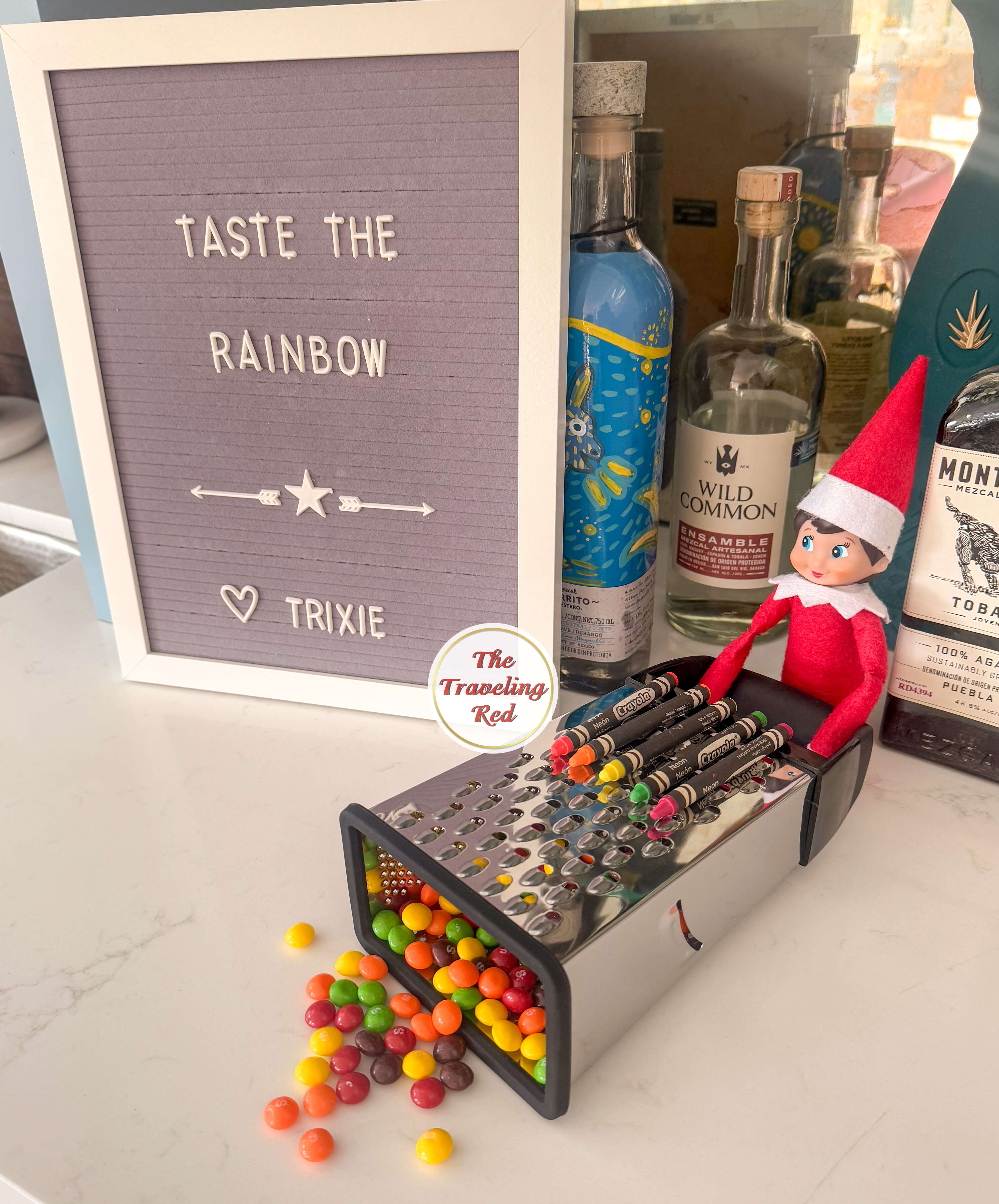 Elf on the Shelf Ideas: Elf is next to a grater with crayons on top and skittles coming out of the bottom. The elf has a sign that says taste the rainbow. Over 100 easy and quick elf on the shelf ideas that are fun. By The Traveling Red
