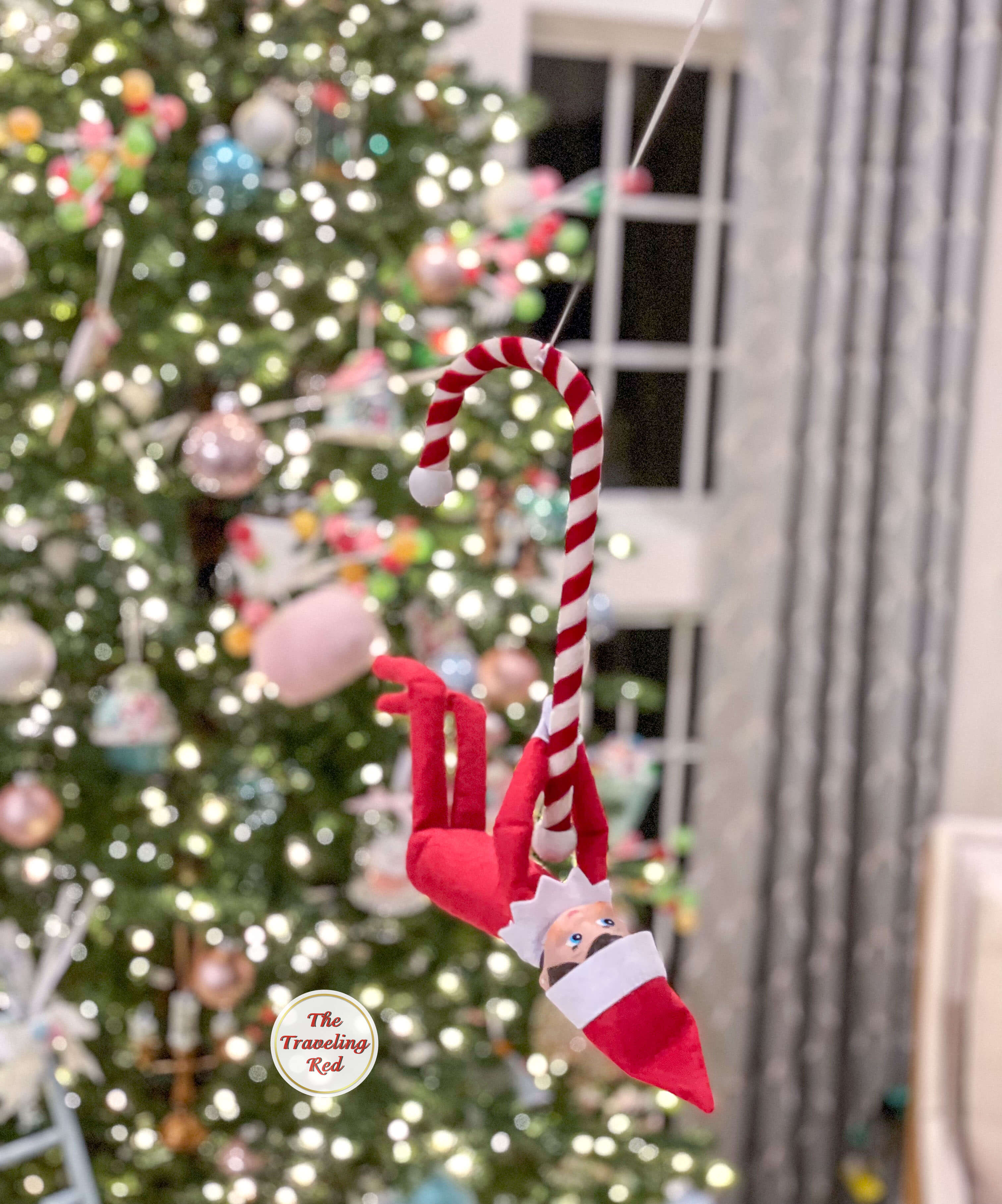 Elf on the Shelf Ideas: Elf is hanging from a zip line she created using a ribbon she attached from the chandelier to the christmas tree and is hanging from a candy cane. Over 100 easy and quick elf on the shelf ideas that are fun. By The Traveling Red