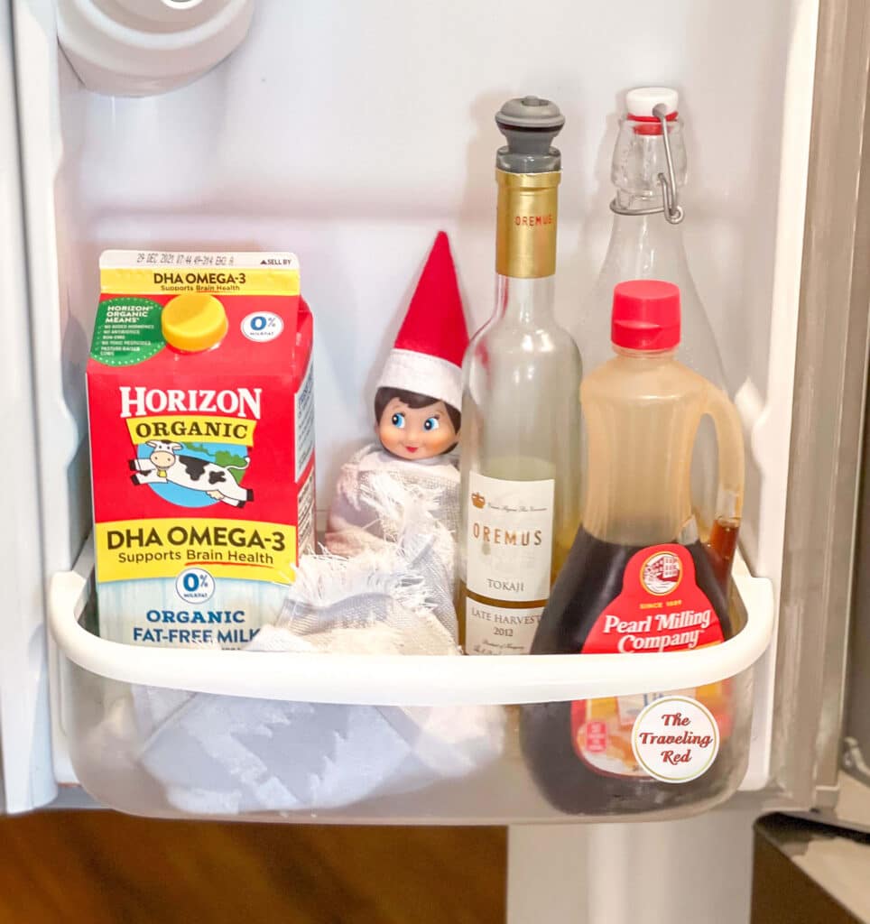 Elf on the Shelf Ideas: Elf is hiding in the fridge door wrapped in a hand towel. Over 100 easy and quick elf on the shelf ideas that are fun. By The Traveling Red
