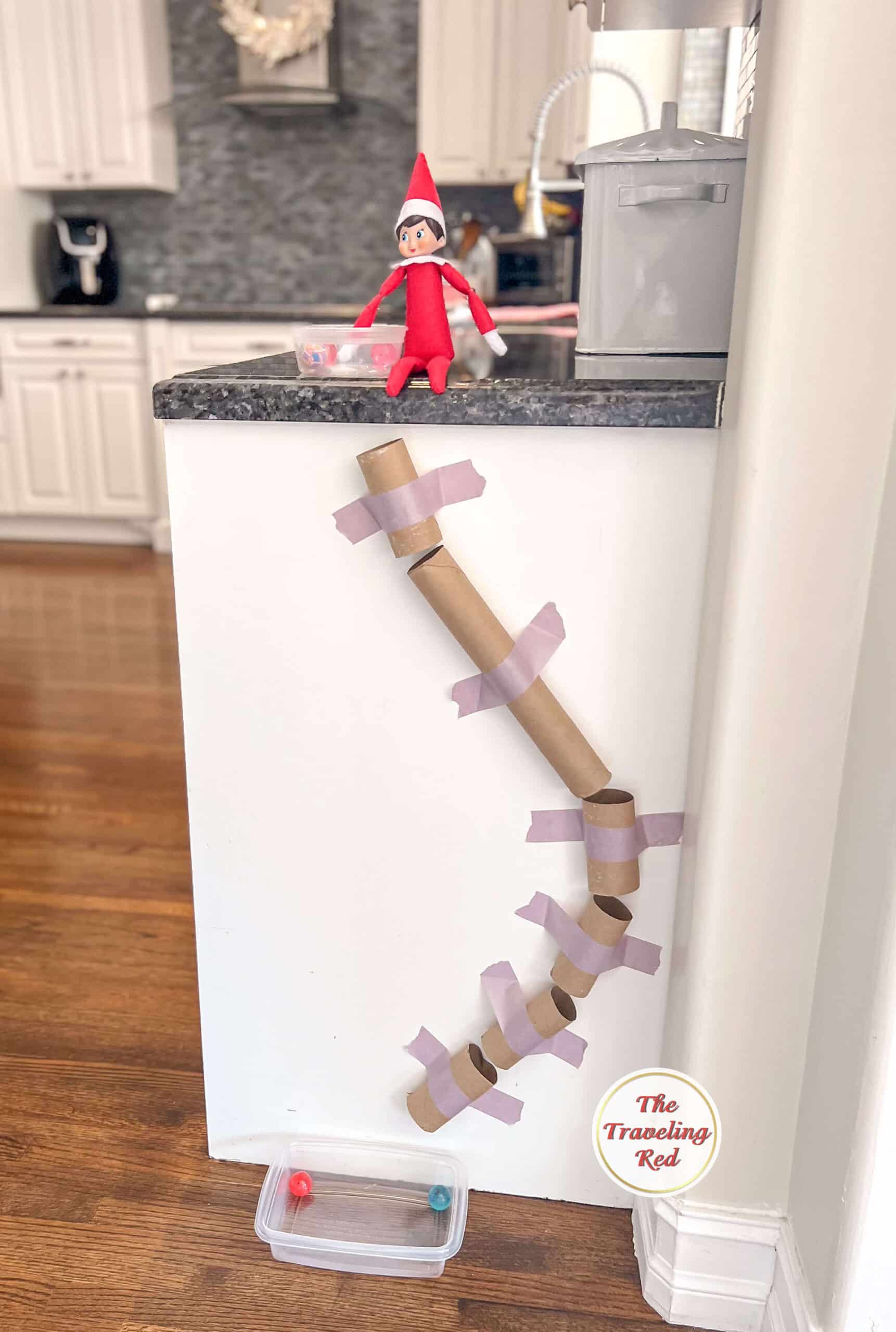 Elf on the Shelf Ideas: Elf used empty toilet paper rolls and paper towel rolls to create a path attached to the kitchen cabinet with painters tape. The path ends at the bottom where is drops into tupperware. There's some small balls to use it. Over 100 easy and quick elf on the shelf ideas that are fun. By The Traveling Red