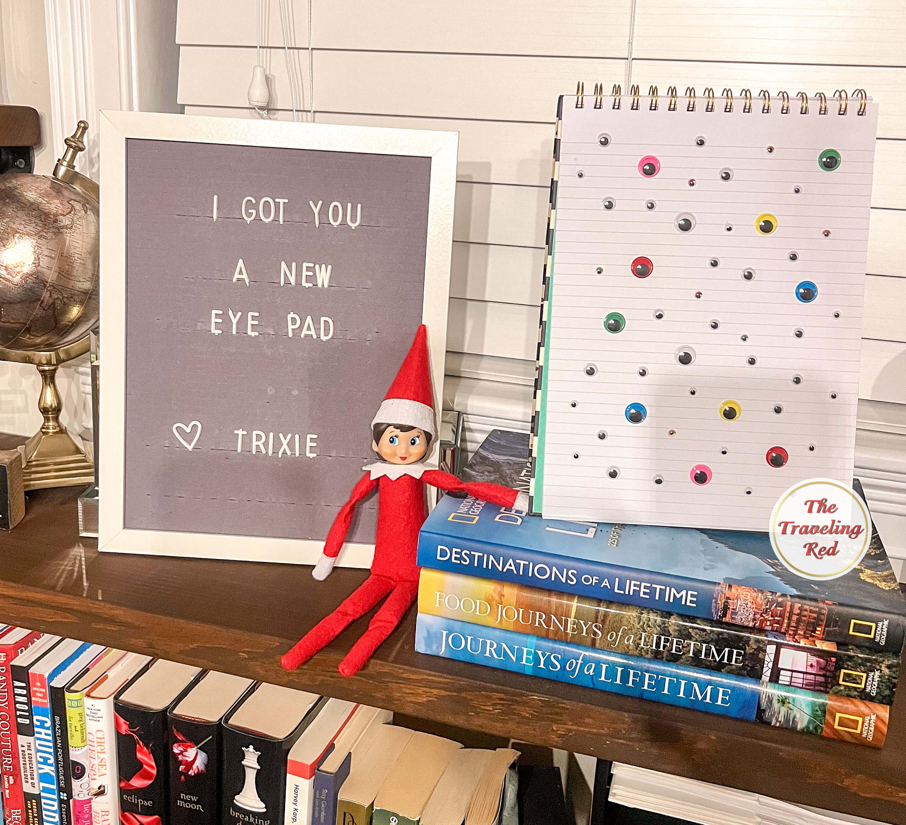 Elf on the Shelf Ideas: Elf is sitting next to a notepad that has the page filled with googly eyes. The message board reads "I got you a new Eye Pad" which is a plain the word iPad. Over 100 easy and quick elf on the shelf ideas that are fun. By The Traveling Red