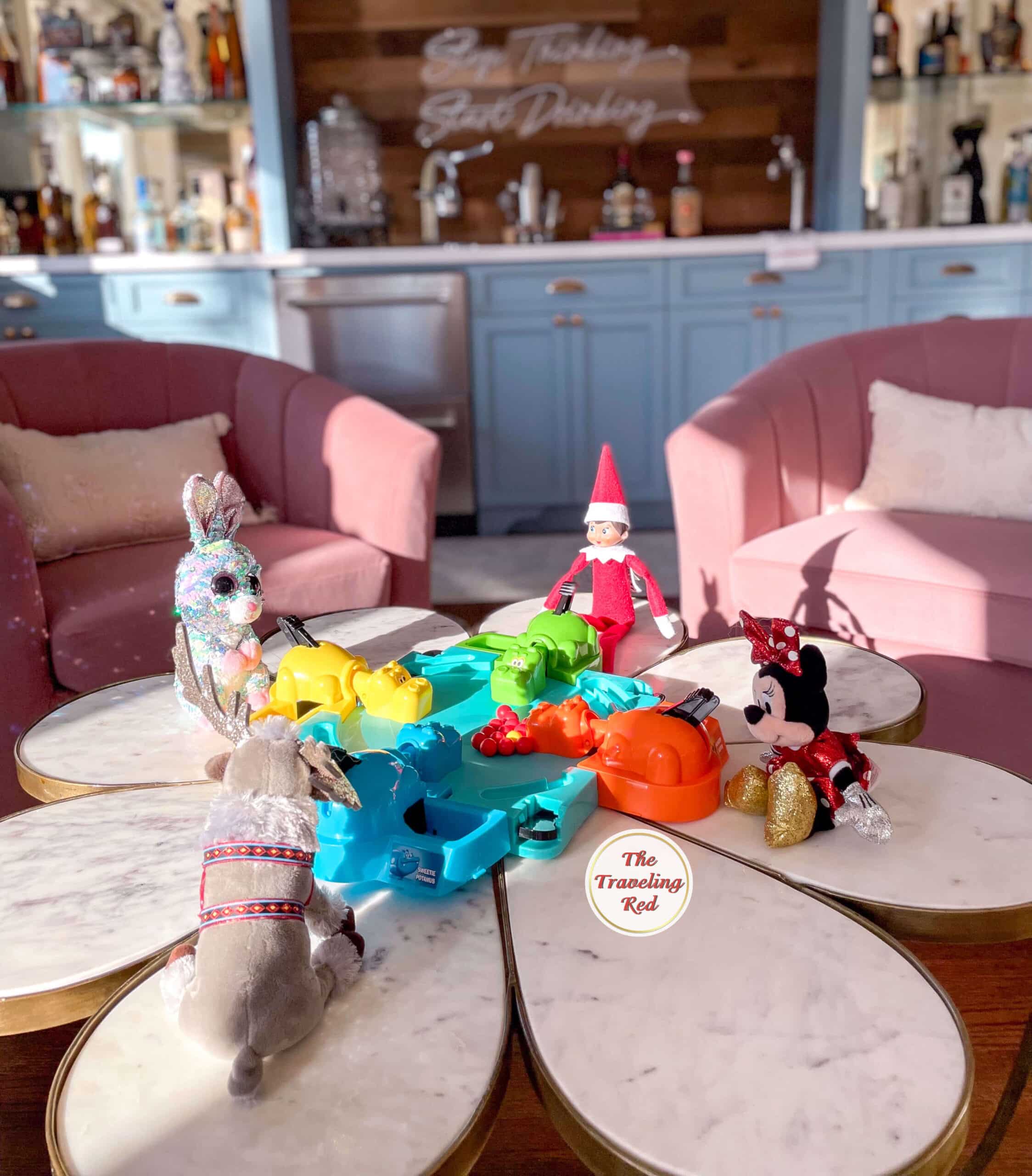Elf on the Shelf Ideas: Elf is sitting on a table with 3 other toys playing hungry hungry hippo. Over 100 easy and quick elf on the shelf ideas that are fun. By The Traveling Red
