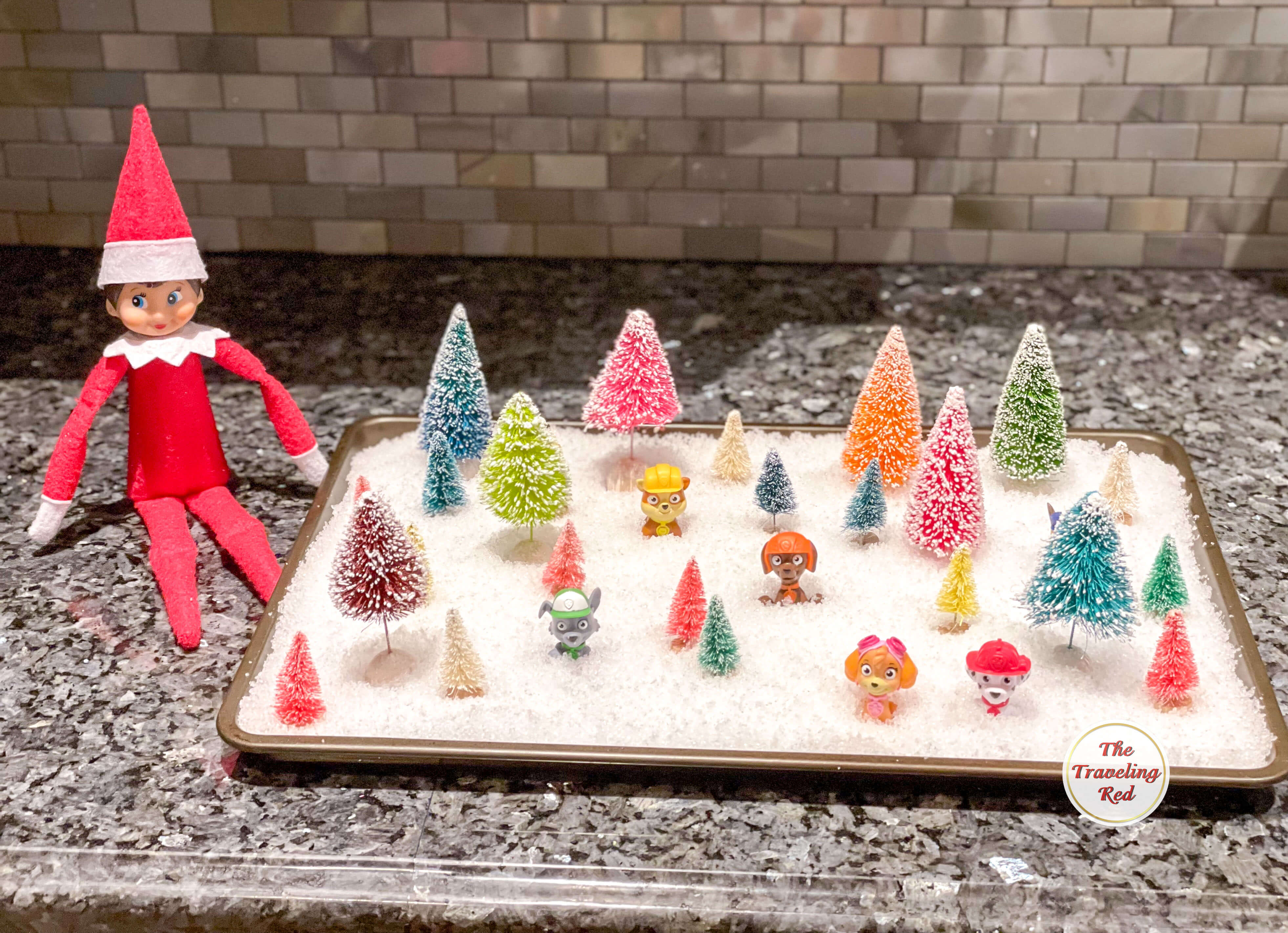 Elf on the Shelf Ideas: Elf is next to a baking sheet covered in snow with trees and little animals in it. Over 100 easy and quick elf on the shelf ideas that are fun. By The Traveling Red