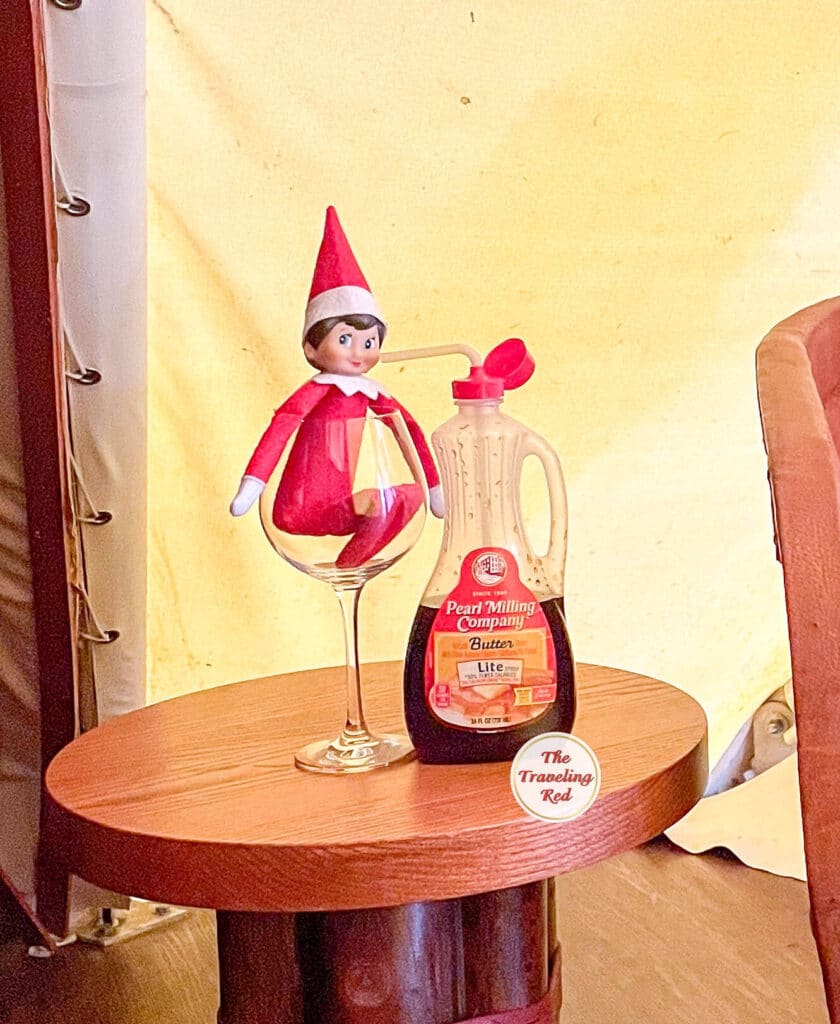 Elf on the Shelf Ideas: Elf is in a wine glass next to a bottle of syrup with a straw in the syrup to drink it. Over 100 easy and quick elf on the shelf ideas that are fun. By The Traveling Red