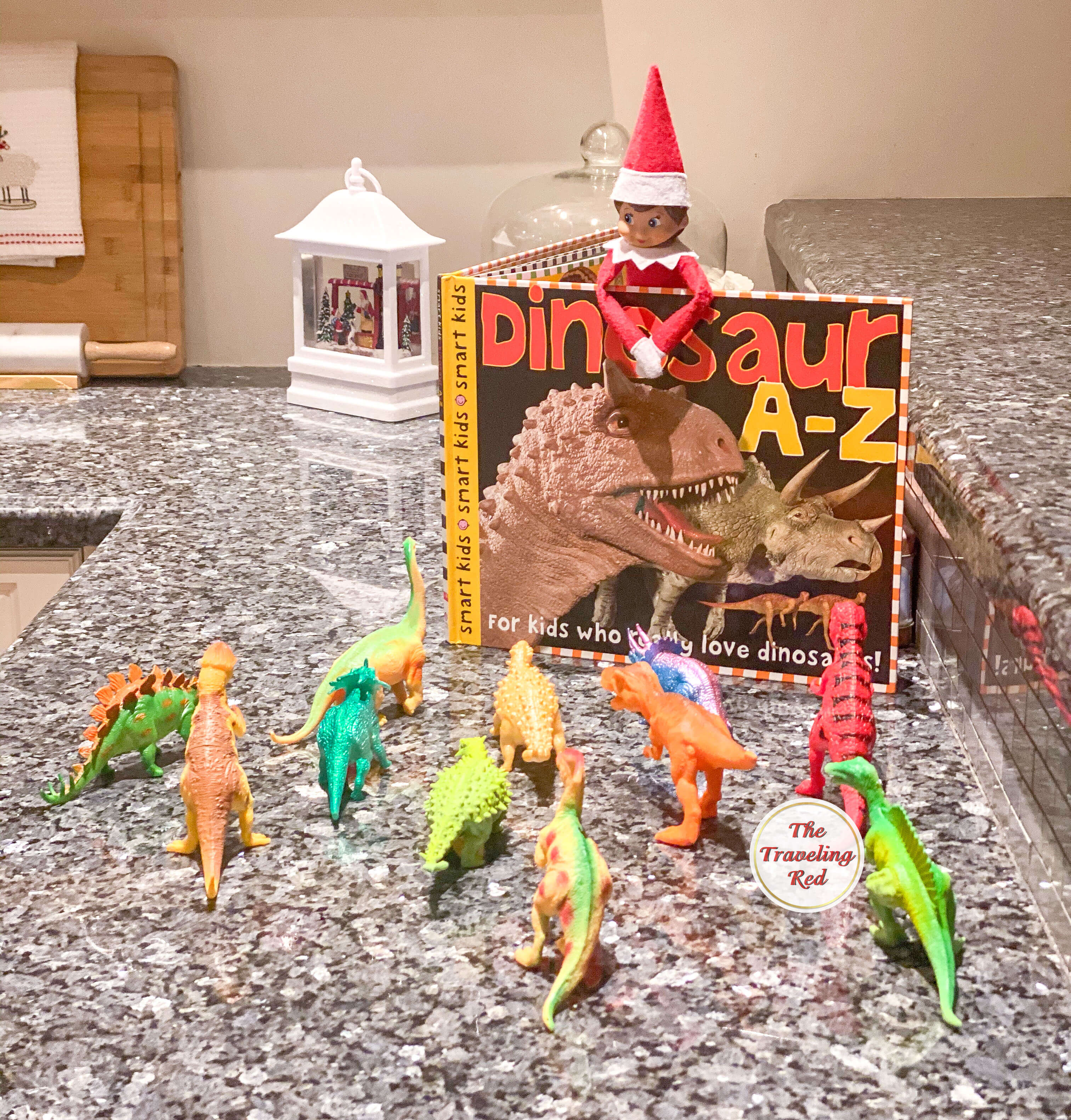 Elf on the Shelf Ideas: Elf is reading a dinosaur book to a lot of little dinosaur figurines. Over 100 easy and quick elf on the shelf ideas that are fun. By The Traveling Red