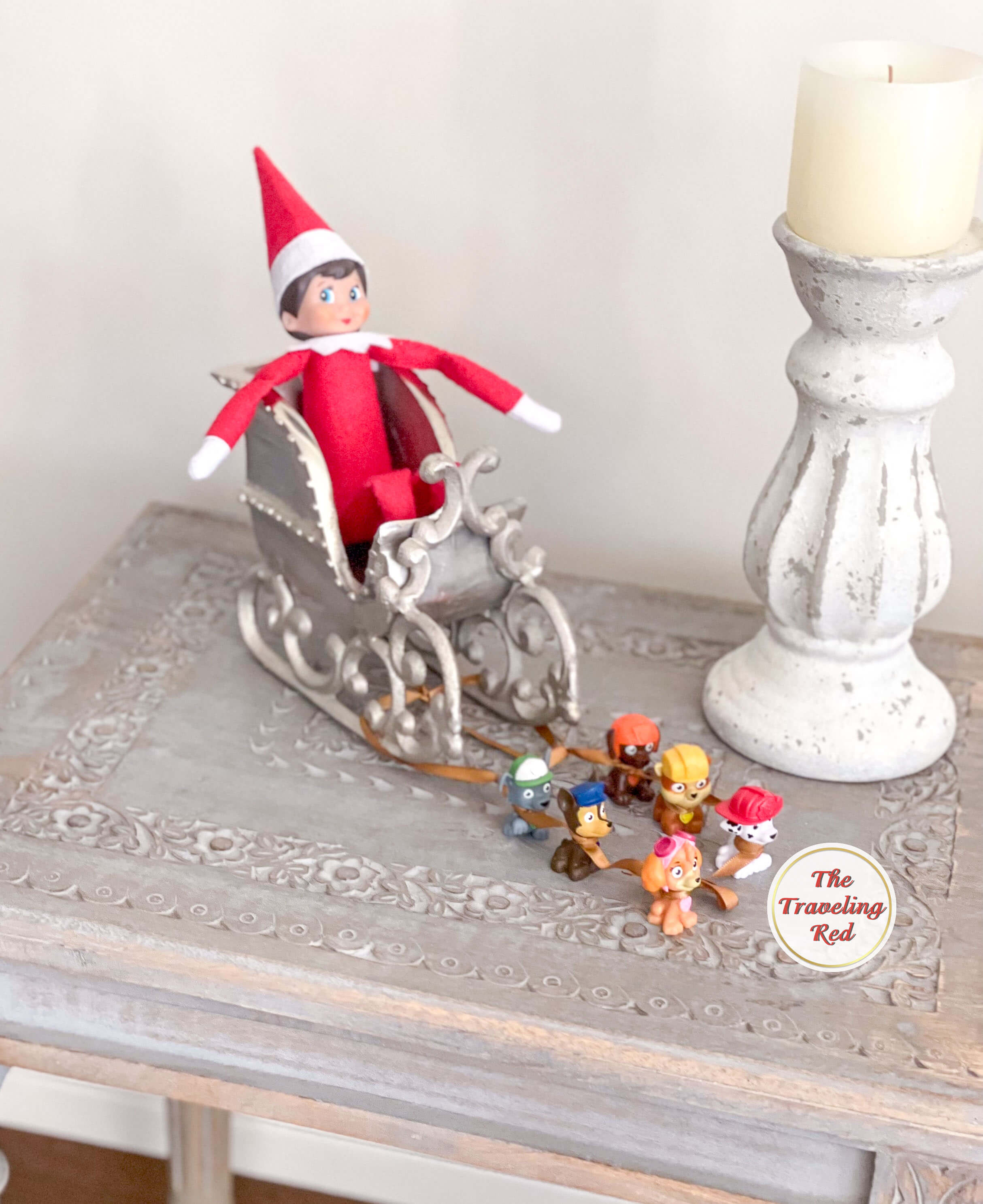 Elf on the Shelf Ideas: Elf is on a decorative sled with had little dog figurines added to the front and they are all tied using a piece of ribbon to create a dog sled. Over 100 easy and quick elf on the shelf ideas that are fun. By The Traveling Red