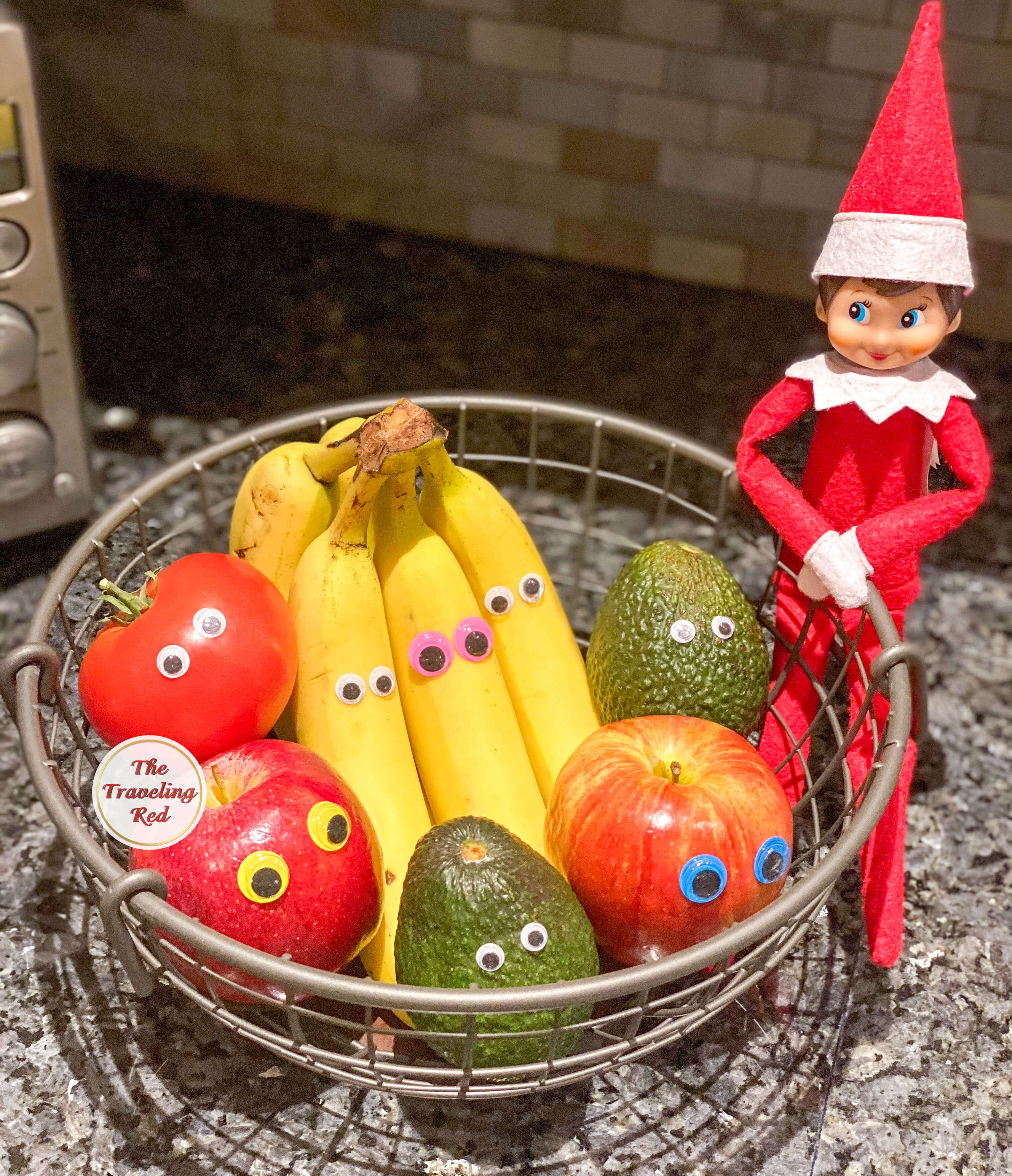 Elf on the Shelf Ideas: Elf added googly eyes to all of the fruit in the fruit basket and is sitting next to it. Over 100 easy and quick elf on the shelf ideas that are fun. By The Traveling Red