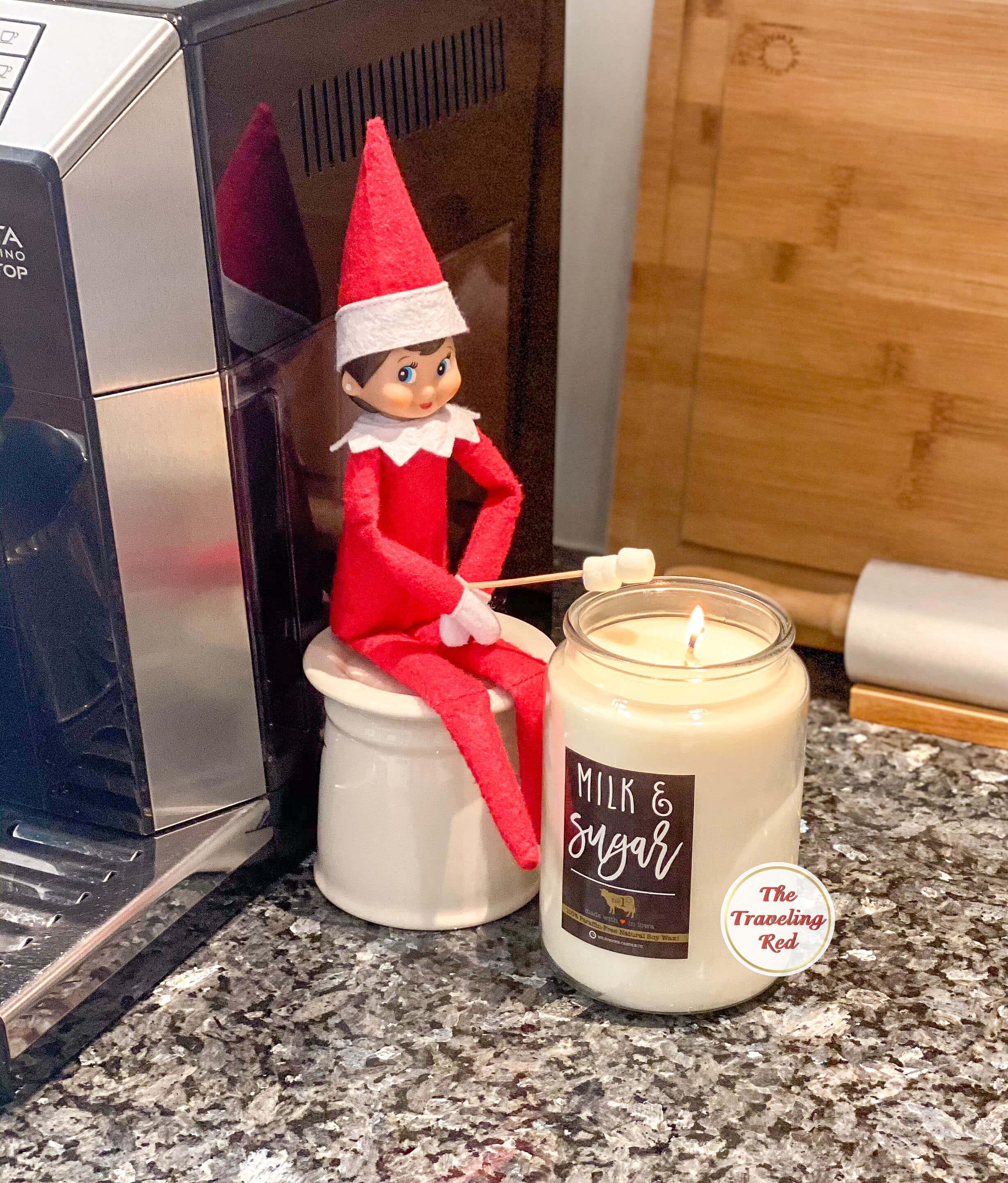 Elf on the Shelf Ideas: Elf is sitting on an upside down coffee mug with a toothpick in hand that has mini marshmallows at the end. She is roasting the marshmallows over a candle to make smores. Over 100 easy and quick elf on the shelf ideas that are fun. By The Traveling Red